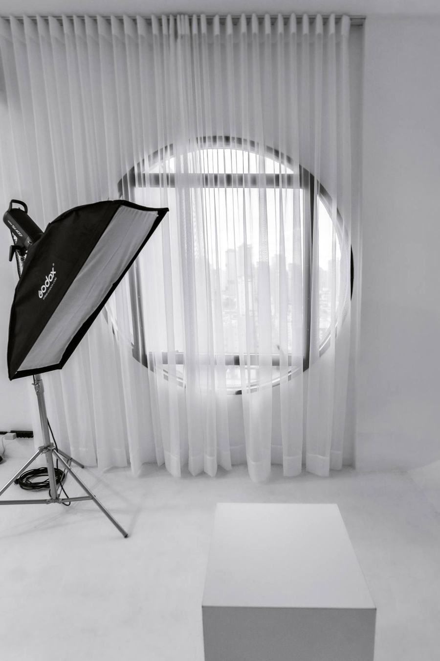 Detalhe do est&uacute;dio da Aliar: janela circular com cortina, softbox montado e cubo branco de apoio